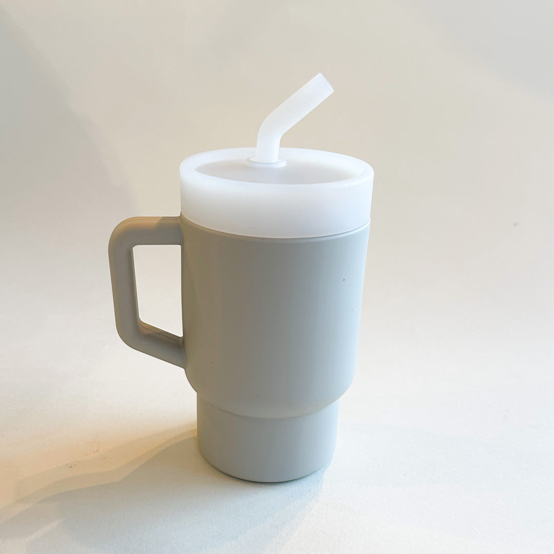 A lightweight 9oz toddler tumbler in soft, premium silicone, designed with a handle for easy grip and portability. The tumbler features a leak-resistant straw with a soft silicone tip, making it perfect for independent sipping. Available in playful colors for kids, it combines functionality with fun for little explorers. Light grey, unisex color for boys and girls.
